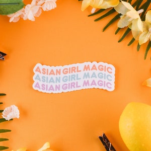Könnte beinhalten: Ein Aufkleber mit dem Text "Asian Girl Magic", der dreimal in einem Regenbogen-Farbverlauf wiederholt wird. Der Aufkleber befindet sich auf einem leuchtend orangenen Hintergrund mit Zitronen und gelben Blumen.