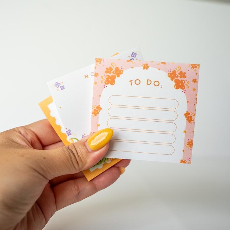 May include: A hand holding a stack of colorful sticky notes. The top note is pink with orange floral designs and the words "TO DO:" above lined spaces. Other notes have floral and geometric designs in orange, purple, and yellow.