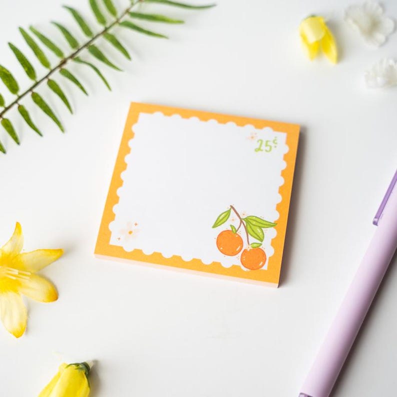Pode incluir: Bloco de notas quadrado laranja com centro branco e borda decorativa. A nota apresenta uma pequena ilustra&ccedil;&atilde;o de laranjas e o texto "25&cent;". Uma caneta roxa e detalhes florais tamb&eacute;m s&atilde;o vis&iacute;veis.