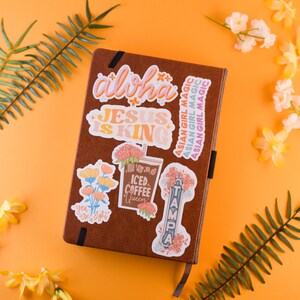 May include: A brown leather journal with colorful stickers on the cover. The stickers feature phrases like "Aloha", "Jesus is King", "Iced Coffee Queen", and "Tampa".