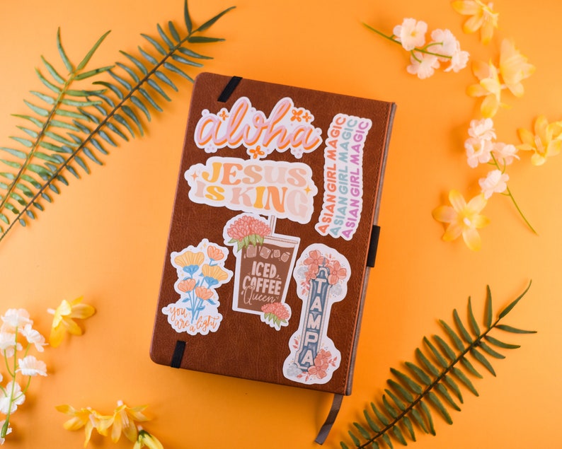 May include: A brown leather journal with colorful stickers on the cover. The stickers feature phrases like "Aloha", "Jesus is King", "Iced Coffee Queen", and "Tampa".