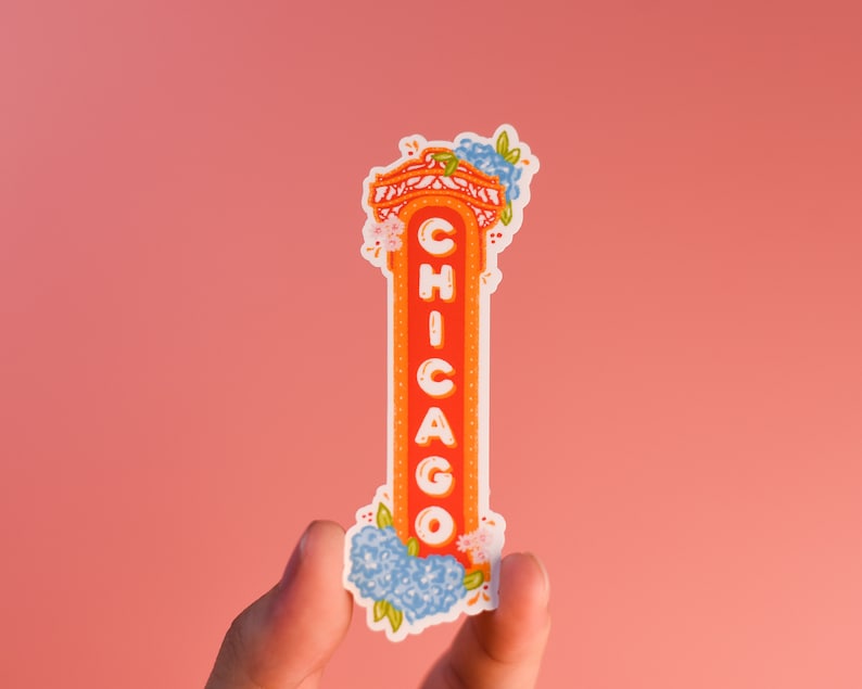May include: A white sticker with a red and orange illustration of a Chicago sign with blue and white flowers on top. The sign says "CHICAGO" in white letters.