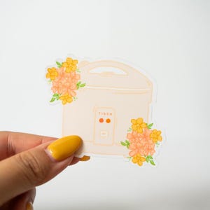 Peut inclure: Autocollant repr&eacute;sentant un cuiseur &agrave; riz p&ecirc;che clair avec le mot "TIGER" imprim&eacute; dessus. L'autocollant est orn&eacute; de motifs floraux orange et jaunes. L'autocollant est tenu par une main avec un ongle peint en jaune.