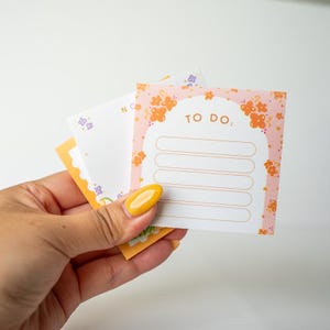 Pode incluir: Uma m&atilde;o segurando uma pilha de notas adesivas coloridas. A nota superior &eacute; rosa com desenhos florais laranja e as palavras "TO DO:" acima de espa&ccedil;os com linhas. Outras notas t&ecirc;m desenhos florais e geom&eacute;tricos em laranja, roxo e amarelo.