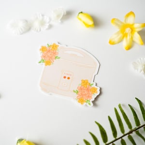 Peut inclure: Un autocollant repr&eacute;sentant un cuiseur &agrave; riz cr&egrave;me avec le mot "TIGER" imprim&eacute; dessus. L'autocollant est orn&eacute; de compositions florales orange et jaune. Le fond comprend des fleurs blanches, des fleurs jaunes et de la foug&egrave;re verte.