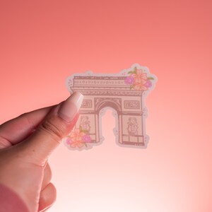 May include: A sticker featuring a detailed illustration of the Arc de Triomphe in Paris, France. The sticker is adorned with pink and orange floral accents. The background is a gradient of pink and peach.