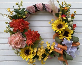 May include: A wreath made of grapevine with a pink ribbon, featuring a variety of artificial flowers, including sunflowers, hydrangeas, geraniums, and small yellow flowers. The wreath is decorated with a green and orange bow.