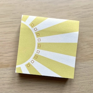 May include: A yellow sticky note with a white sun design and a brown border. The sun has a circle of small squares around it.