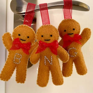 May include: Three felt gingerbread man decorations with red ribbon bows and hanging loops. Each gingerbread man has a stitched outline, black eyes, and a letter stitched on the front. The ornaments are suspended from a silver door handle.
