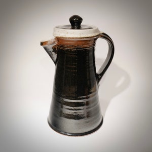 May include: A handmade ceramic coffee pot with a black glaze. The pot has a white rim, a brown band, and a black lid. It features a handle and a spout for pouring. The design is simple and elegant, suitable for kitchen decor.