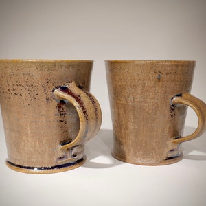 May include: Two handmade ceramic mugs with a speckled, earthy brown glaze. Each mug features a curved handle and a dark blue accent at the base and handle attachment. The mugs have a slightly tapered shape.