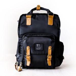 May include: Black backpack with brown leather accents and buckles. The backpack has a large main compartment and a smaller front pocket with the logo "Too Loot Australia".