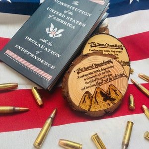 May include: A close-up of a wooden coaster with the text "The Second Amendment" and a stylized image of mountains and an eagle. The coaster is on top of a book titled "The Constitution of the United States of America with the Declaration of Independence". There are several gold-colored bullets scattered around the coaster and book.