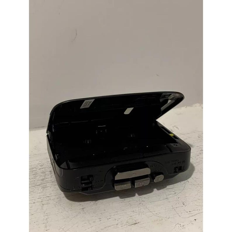 May include: Black cassette player with the lid open. The player has a silver button on the front and the text "SONY" is visible on the side.