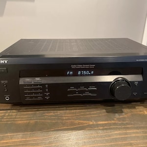 Pode incluir: Receptor estéreo Sony preto com um visor digital que mostra "FM 8750.A". O receptor tem uma variedade de botões e botões para controlar o áudio e o vídeo.