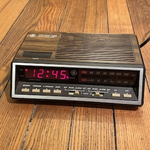 Puede incluir: Un radio despertador vintage GE marrón y plateado con una pantalla digital que muestra las 12:45. El radio despertador tiene un acabado de madera y cuenta con bandas de radio AM y FM, dos horas de despertador y un temporizador de apagado.