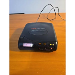 Può includere: Lettore CD Sony Car Discman nero con display digitale che mostra le informazioni sulla traccia. Il dispositivo ha controlli di riproduzione, pausa, salto e volume. Un filo nero si estende dalla parte superiore dell'unità.