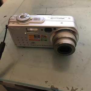 Op de afbeelding: Een zilveren Sony Cyber-shot 3,2 megapixel digitale camera met een zoomlens. De camera heeft een zwarte band en een zwarte lensdop. De camera heeft een aantal knoppen en bedieningselementen, waaronder een aan/uit-knop, een zoomknop en een sluiterknop.