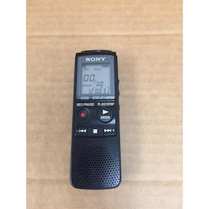 May include: A black Sony digital voice recorder featuring a rectangular LCD screen displaying numerical data, a circular record button with a red dot, a play/stop button, navigation controls, and a perforated speaker grille at the bottom.