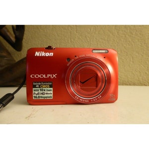 May include: A red Nikon Coolpix digital camera with a 10x wide optical zoom lens. The camera has black and silver trim and features a back-illuminated CMOS sensor, full HD movie recording, and 16.0 megapixels.