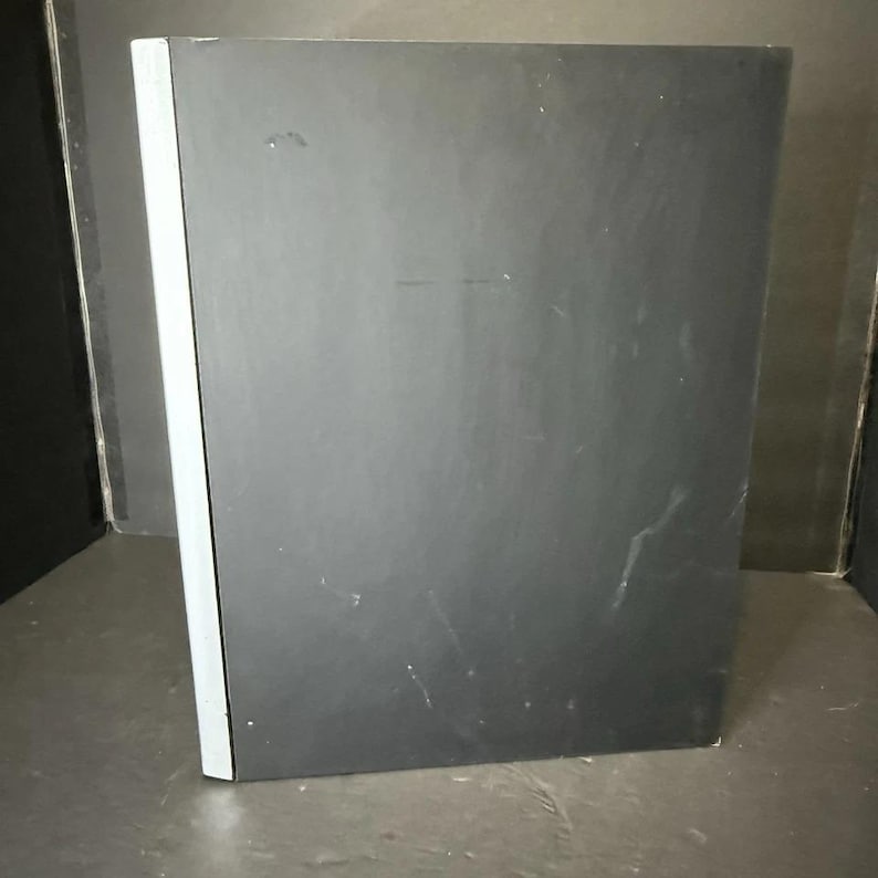 May include: A black book with a white spine. The book is rectangular and appears to be closed. The cover has some minor scratches and marks. The book is set against a dark background.