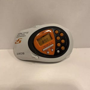 Może przedstawiać: Biało-pomarańczowe radio Sony z wyświetlaczem cyfrowym pokazującym 87,7. Radio ma wiele przycisków, w tym preselekcję, pasmo, tryb, strojenie/ustawianie czasu i enter. Radio ma również gniazdo słuchawkowe i przycisk hold.