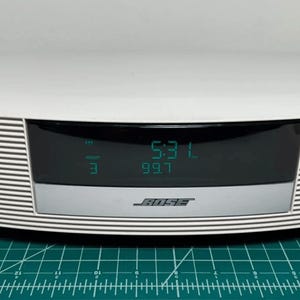 May include: White Bose radio with a digital display showing the time and radio station. The radio has a curved design with horizontal black stripes on the sides. The Bose logo is visible on the front.