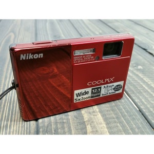 May include: A red Nikon Coolpix digital camera with a 3.5 inch OLED touch screen and a 12.1 megapixel camera. The camera has a 5x zoom lens and a wide-angle lens.