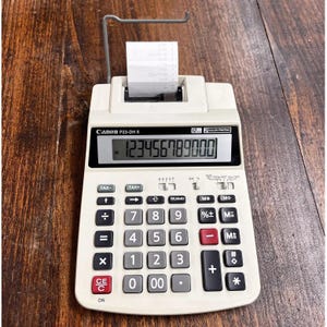 Puede incluir: Una calculadora Canon P23-DH II blanca con una impresora integrada. La calculadora tiene una pantalla grande que muestra el número 123456789000. La calculadora tiene una variedad de botones, incluidos números, operadores y funciones.