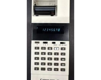 May include: A white Canon TP-8 pocket printer with a black and white calculator keypad and a small printer roll on top. The display shows the number 123456.78.
