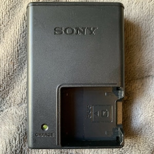 May include: Black Sony battery charger with a green charging light and a slot for a battery. The charger has the word "CHARGE" printed on the front.