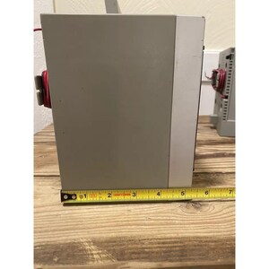 May include: A gray metal electrical box with a red wire coming out of the top. The box is sitting on a wooden surface and is being measured with a yellow tape measure. The tape measure reads 6 inches.
