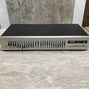 Puede incluir: Un ecualizador de frecuencia estéreo de 12 bandas Realistic plateado con botones negros y una parte superior negra. El ecualizador tiene una placa frontal plateada con el texto "Realistic Twelve Band Stereo Frequency Equalizer" impreso en él. El ecualizador tiene una serie de deslizadores para ajustar las bandas de frecuencia. El ecualizador tiene un botón de encendido y una serie de conectores de entrada y salida.