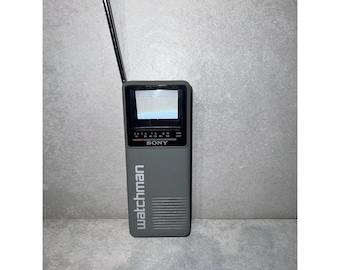 May include: A gray Sony Watchman portable television with a black and white screen and a telescopic antenna. The television has a silver control panel with buttons and a volume knob.