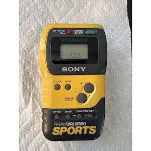 May include: A yellow and black Sony Sports FM/AM Walkman radio with a digital clock display. The radio has several buttons, including clock, mode, memory preset, enter, band, and tune/time set.