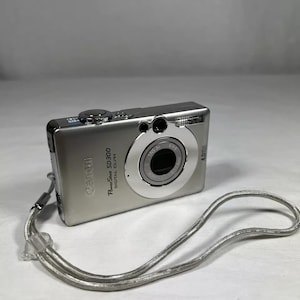 May include: A silver Canon PowerShot SD300 digital camera with a black lens and a silver wrist strap.