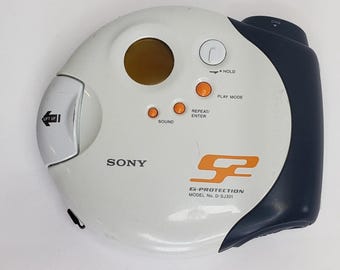 May include: A white and navy blue Sony portable CD player. The player has orange buttons for sound, play mode, and repeat/enter. The model number is D-SJ301. The player has a silver lift-up door and a dark brown circular window.