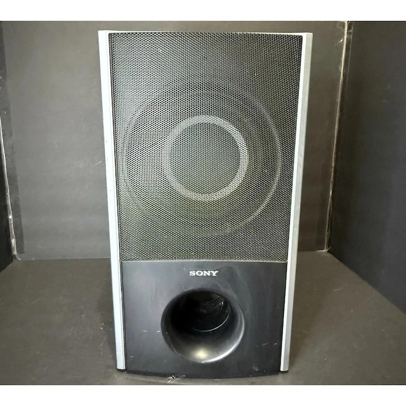 May include: A black Sony subwoofer speaker with a silver frame. The speaker has a mesh front with a large circular design in the center. The bottom section is solid black with a circular port. The Sony logo is visible.