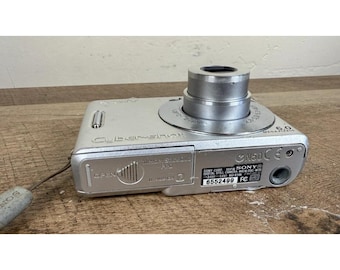 May include: Silver Sony Cybershot digital camera with a retractable lens and a built-in flash. The camera has a 6.0-megapixel resolution and a small LCD screen. The camera is on a wooden surface.