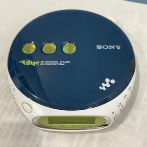 Puede incluir: Un reproductor de CD Sony Walkman D-EJ360 azul marino y blanco. El reproductor de CD portátil tiene una pantalla digital que muestra información de pista y tiempo. El dispositivo presenta un borde blanco, tres botones iluminados y el logotipo de Sony. También es visible el texto "G-PROTECTION".