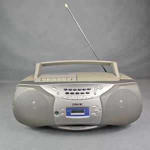 May include: A vintage Sony CFD-Z330 boombox in silver and beige. The radio cassette CD player has a handle, antenna, and a digital display. The front features two speakers and the text "MEGA BASS PORT".