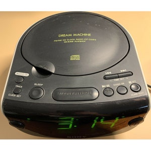 Puede incluir: Radio despertador Sony Dream Machine FM/AM CD ICF-CD815 negro con una pantalla digital verde que muestra 3:14. La radio tiene un reproductor de CD, temporizador de apagado y funciones de despertador.