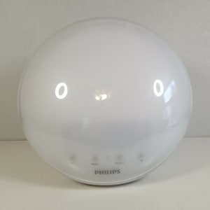 Puede incluir: Luz de despertador Philips redonda blanca con una luz blanca que emite desde la parte superior. La luz tiene tres botones etiquetados como "Menú", "Seleccionar" y "+".