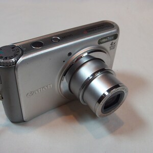 May include: Silver Canon digital camera with a retractable lens. The camera has a 4x optical zoom and a mode dial on the top. A wrist strap is attached to the side of the camera.