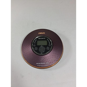 Puede incluir: Un reproductor de CD Sony Walkman burdeos, modelo D-NE320, con el texto "PSYC" y "Sony" visibles. El dispositivo tiene un diseño circular con una superficie perforada y un borde naranja. Cuenta con una pantalla digital y botones de control.