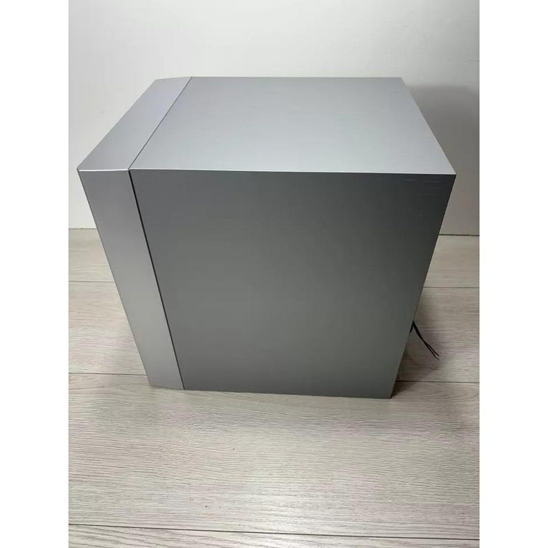 Pu&ograve; includere: Un altoparlante rettangolare di colore argento con una superficie liscia e piatta. L'altoparlante &egrave; appoggiato su un pavimento in legno chiaro.