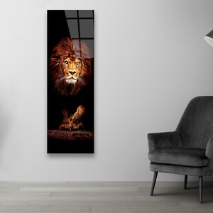 May include: A vertical rectangular wall art print featuring a majestic lion. The lion's face and mane are a warm brown color, set against a dark background. The artwork is displayed on a white wall, with a black chair and lamp in the room.