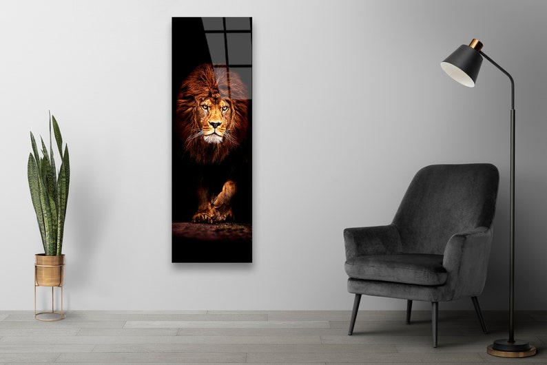May include: A vertical rectangular wall art print featuring a majestic lion. The lion's face and mane are a warm brown color, set against a dark background. The artwork is displayed on a white wall, with a black chair and lamp in the room.