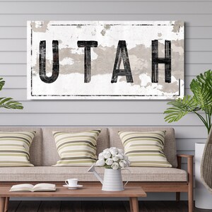 May include: A distressed white sign with black lettering that reads "UTAH". The sign is hanging on a white wood wall above a couch with striped pillows.
