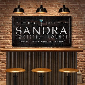 May include: A custom-made sign for a cocktail lounge, featuring the name "SANDRA" in large white letters. The sign includes the text "COCKTAIL LOUNGE" and "Proudly serving whatever you bring" on a dark background, with a martini glass graphic.